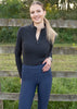Woman wearing a black top and blue classy equestrians high-rise riding tights standing in front of a wooden fence with greenery in the background
