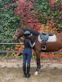 Person standing next to a brown horse with a saddle in an outdoor setting with green and red foliage.